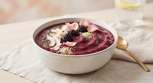 An elegant high-fashion close-up of a smoothie bowl with a deep ruby-violet base made of wild blackberries, açaí, and Peruvian camu camu. Artfully topped with hydrated chia seeds, coconut ribbons, and edible blush rose petals. The ceramic bowl is illuminated by soft high-key studio lighting with warm blush ambient background tones, showing the velvet texture of the antioxidant smoothie.