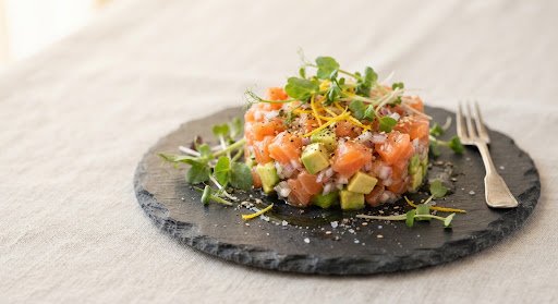 A refined gourmet culinary presentation of wild Alaskan salmon tartare served on a handcrafted slate stone dish. Precision cubes of vivid pink wild salmon, creamy ripe Hass avocado, fresh Japanese yuzu zest, and delicate microgreens. Drizzled with cold-pressed extra virgin olive oil under warm gentle side lighting with a soft blurred neutral linen backdrop.